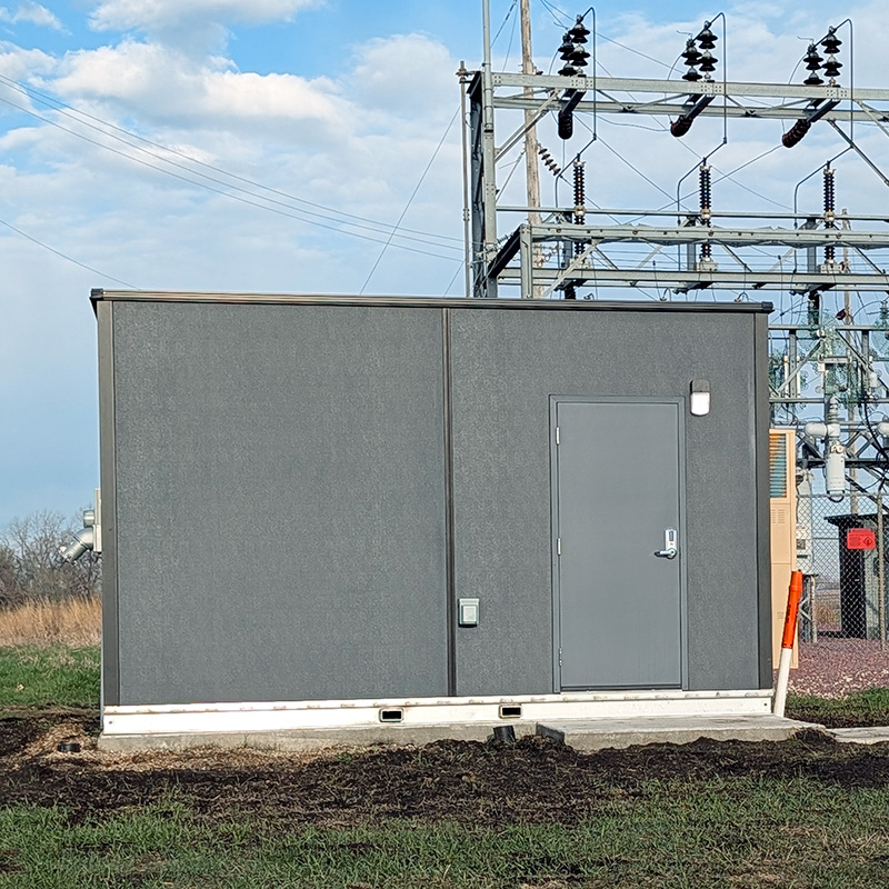 10ft-by-16ft-shelter-installed-cropped Exterior view of a prefabricated utility control building installed at an electrical substation, featuring a secure steel access door and durable weather-resistant construction designed to protect critical power distribution, automation, communications, and substation control equipment.
