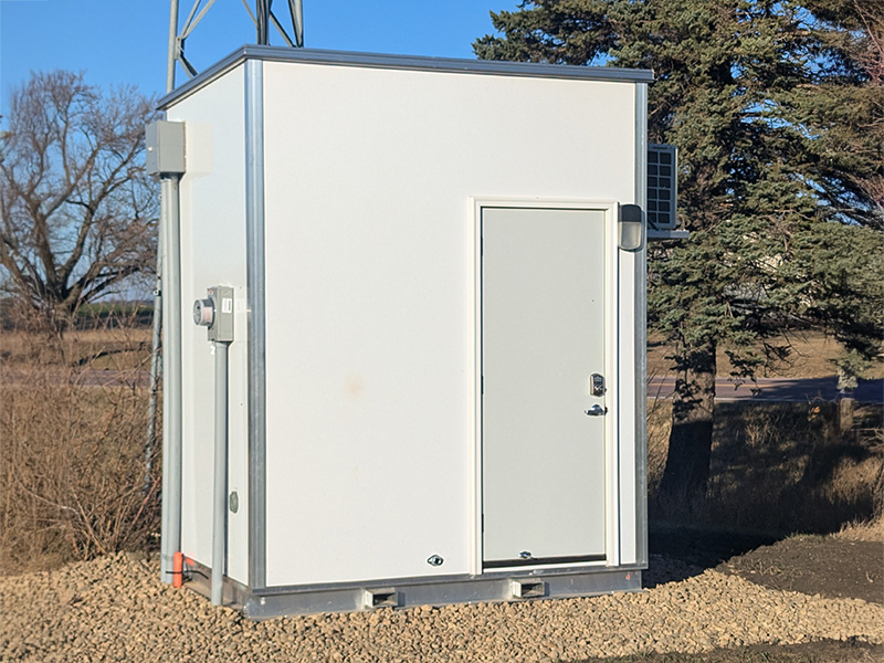 Compact prefabricated utility shelter installed at a communications or utility site, featuring a secure steel access door, exterior electrical service equipment, and climate-control components for protecting critical monitoring, control, and telecommunications systems.